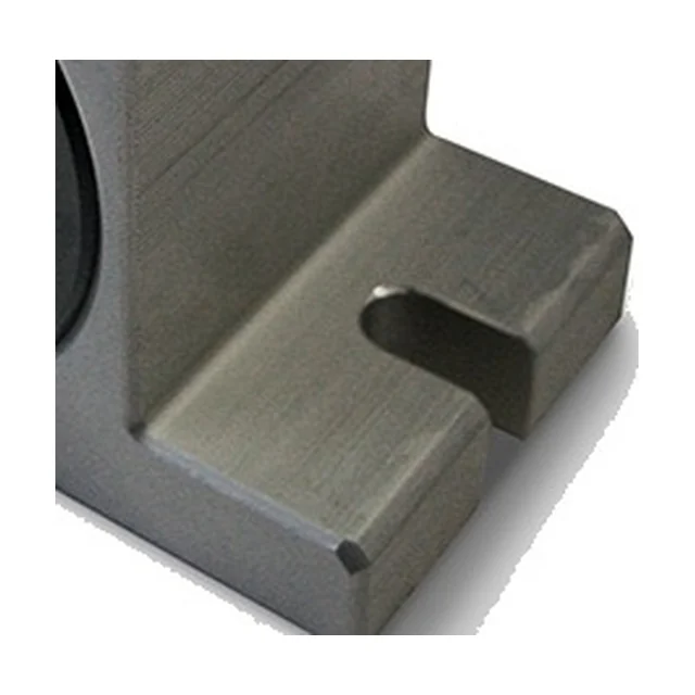 Close-up of one end of the foot on a turbine vibrator, seen from above at an angle. An elongated slot with a rounded end lies in the top face of the foot and opens out towards its edge. On the left is the edge of the black cover. The material is untreated aluminium with visible machining marks.