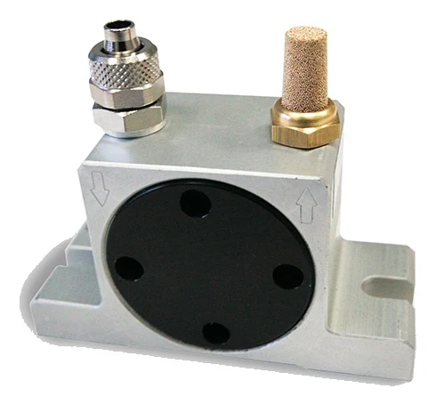 A turbine vibrator seen from the front at an angle: a light aluminium body, clearly wider than it is tall, with a large round black cover in the middle of the side. Four through holes sit near the rim of the cover. On top are two connections: on the left a metal push-in fitting, on the right a brass nut carrying a sintered bronze silencer. An arrow is cast into the front face under each connection: on the left an arrow pointing down, on the right an arrow pointing up. At the bottom the body meets a wide foot with an open slot in the end.