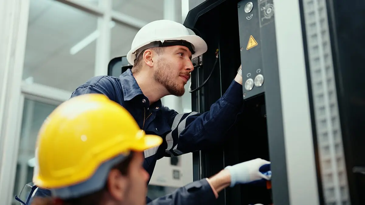 A technician in a hard hat adjusting instruments on a control cabinet while a colleague looks on
