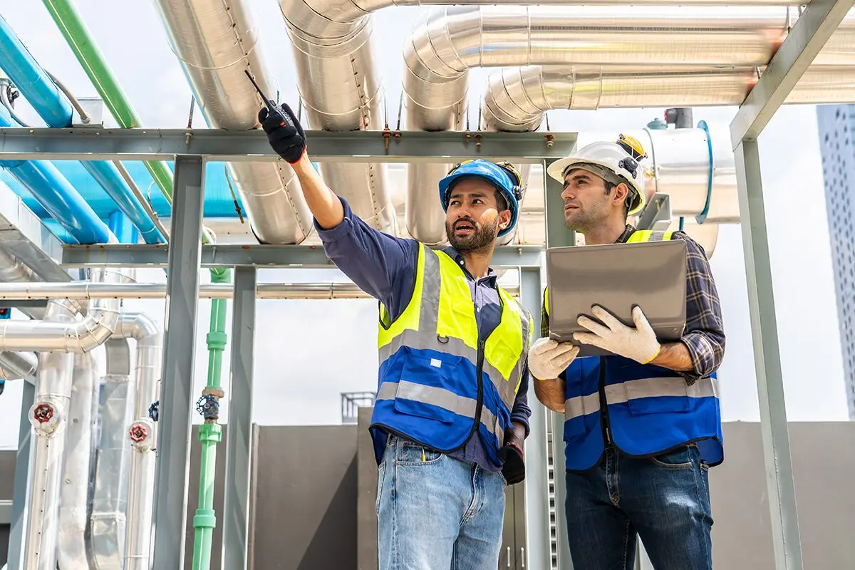 Two technicians reviewing overhead process piping, one with a radio, the other with a laptop